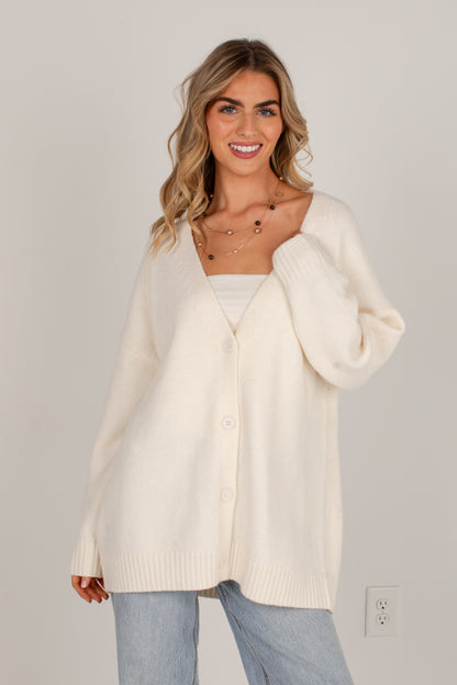 A woman wearing an oversized cream-colored knit cardigan sweater over a white top and light blue jeans, standing against a plain light gray wall.