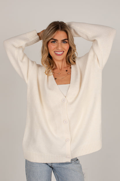 A woman wearing an oversized cream-colored knit cardigan sweater with buttons, paired with light blue jeans, posing with her hands behind her head against a solid light gray background.