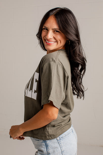 A woman with long dark hair smiling and looking back over her shoulder, wearing an oversized olive green graphic t-shirt with white text and light-wash blue jeans, standing against a light gray brick wall.