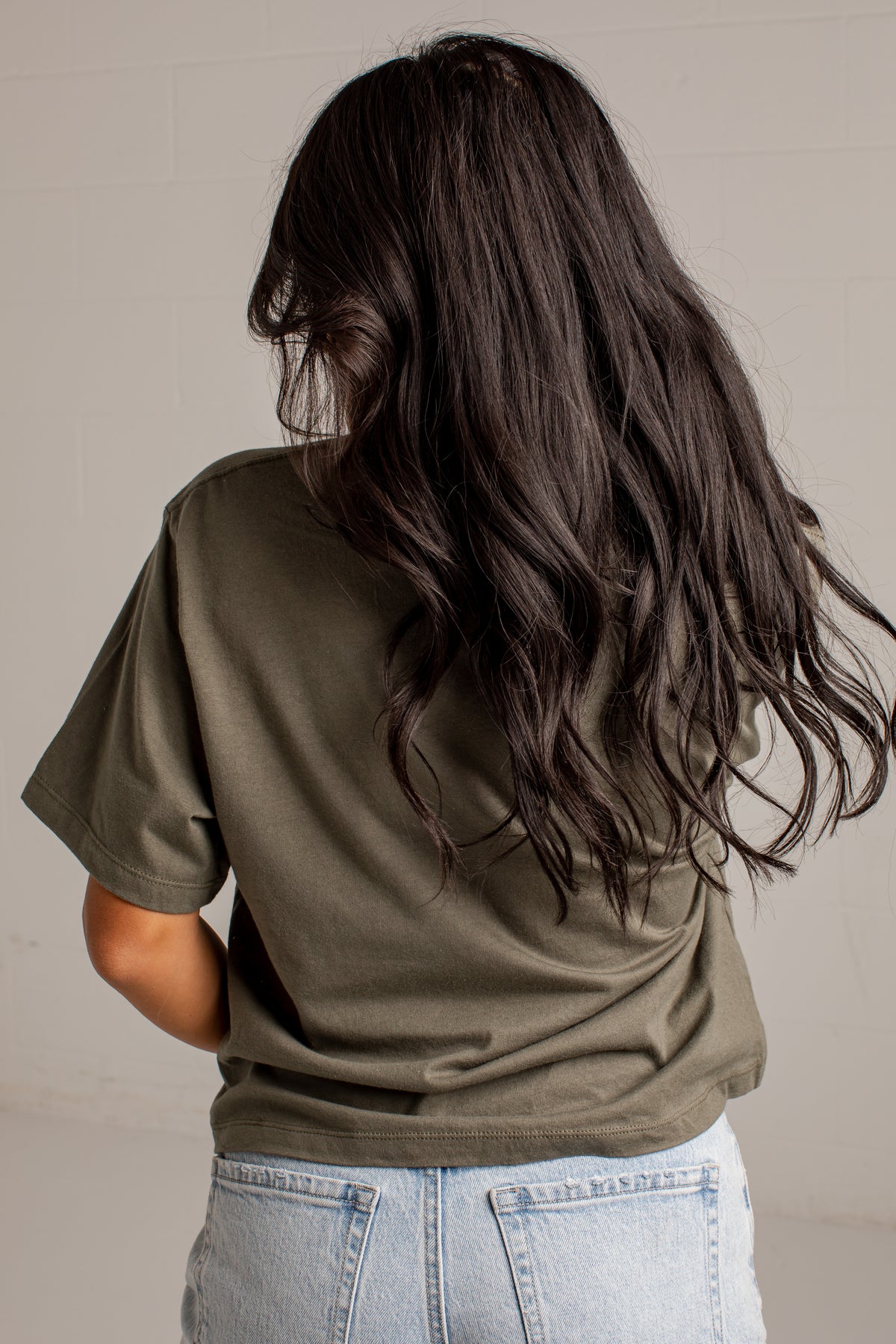 A woman viewed from behind wearing an olive green short-sleeve t-shirt and light wash blue jeans, with her long dark hair blowing in the wind against a light gray wall.