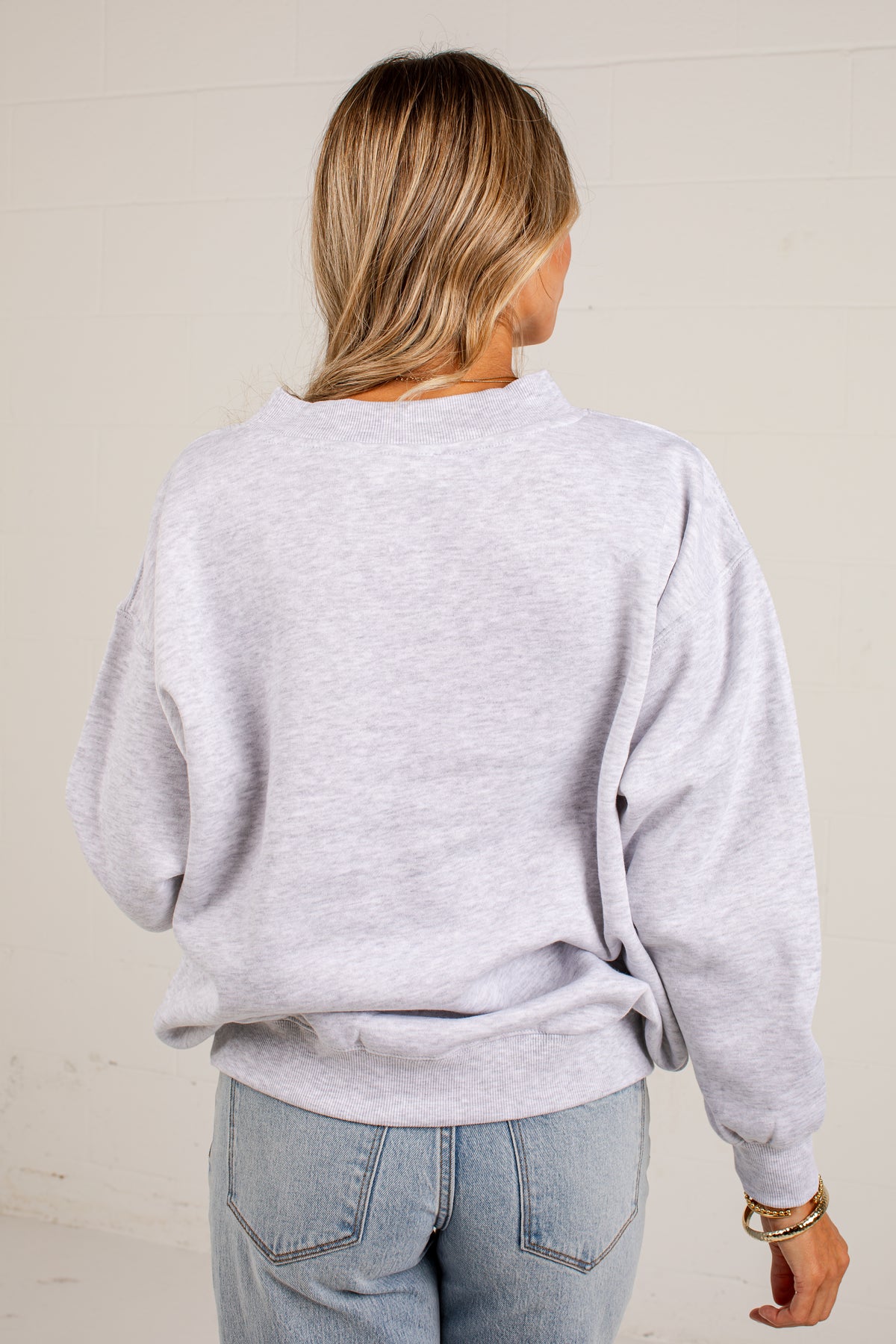 A woman viewed from behind wearing a light heather gray crewneck sweatshirt and light blue jeans, standing against a plain off-white wall.