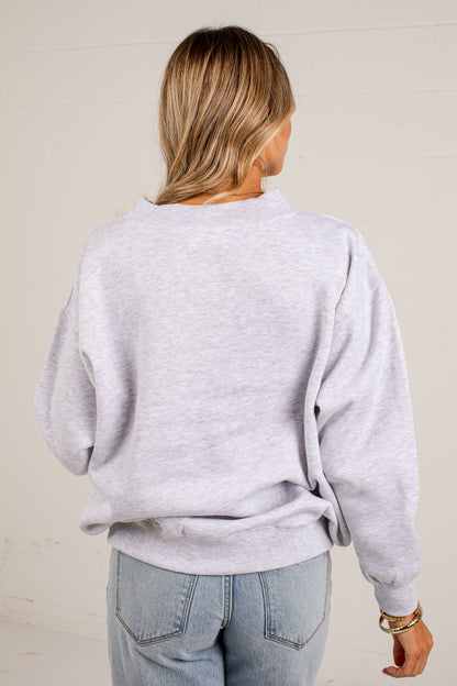 A woman viewed from behind wearing a light heather gray crewneck sweatshirt and light blue jeans, standing against a plain off-white wall.