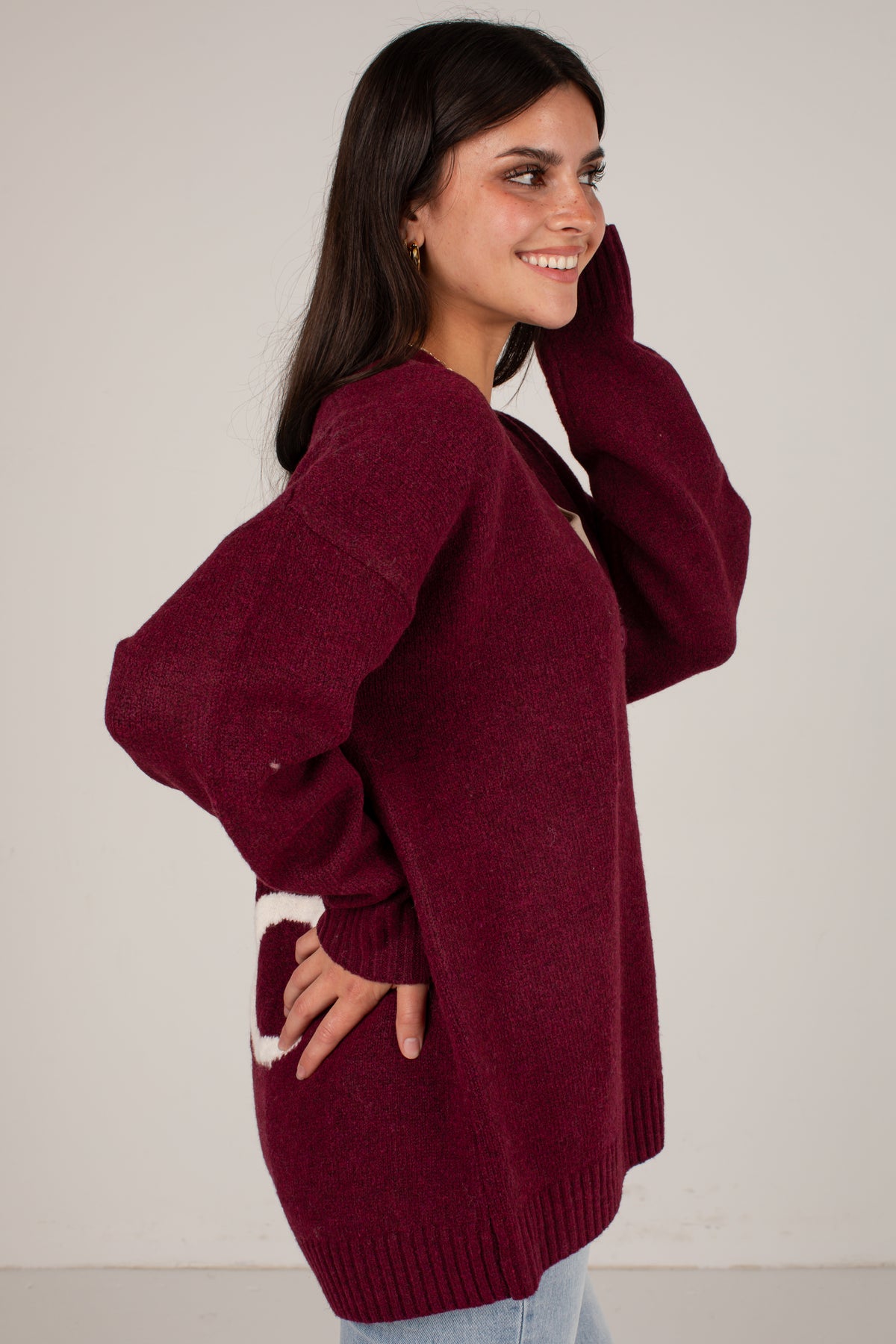 A woman wearing an oversized maroon knit sweater with a white graphic detail on the lower front, posing against a plain off-white background.