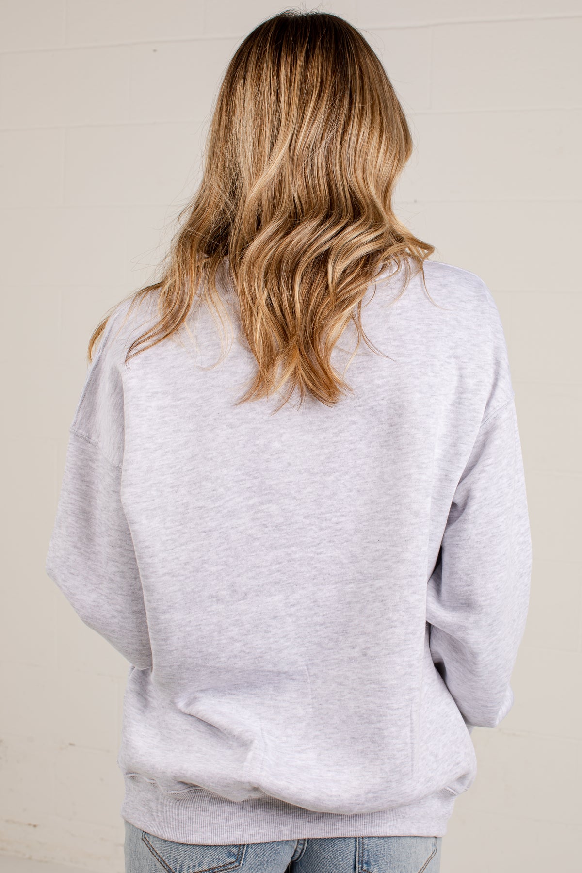 A woman viewed from behind wearing an oversized light gray heather sweatshirt and light blue jeans, standing against a plain off-white wall.