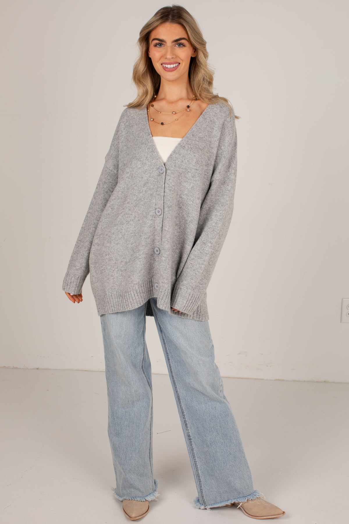 A woman modeling an oversized light gray knit cardigan over a white top, paired with light-wash wide-leg jeans and beige flats, against a plain off-white studio background.