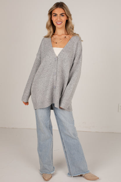 A woman modeling an oversized light gray knit cardigan over a white top, paired with light-wash wide-leg jeans and beige flats, against a plain off-white studio background.