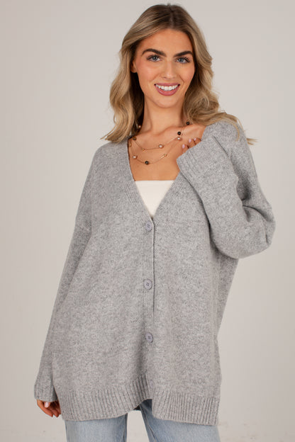 A woman wearing an oversized light gray knit cardigan with a V-neck and button-front closure, layered over a white top and light blue jeans, against a plain light gray studio background.