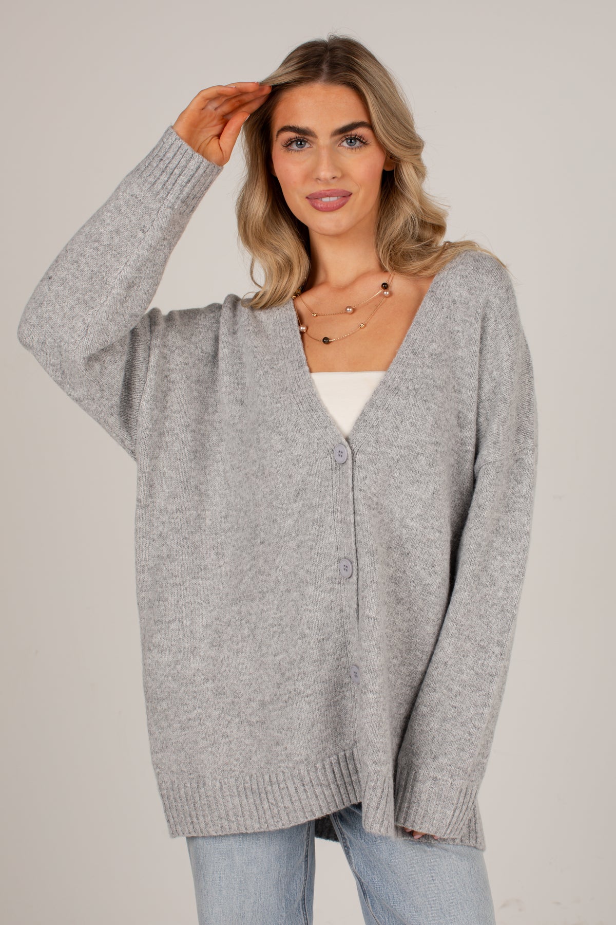 A woman wearing a light gray oversized knit cardigan over a white top and light blue jeans, standing against a plain light gray studio background.