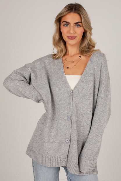 A woman wearing a light gray knit cardigan with a V-neck and front buttons, layered over a white top and light blue jeans, photographed against a plain light gray studio background.