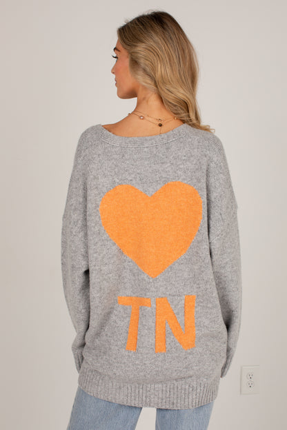 A woman shown from the back wearing a heather gray knit sweater with a large orange heart and the letters &