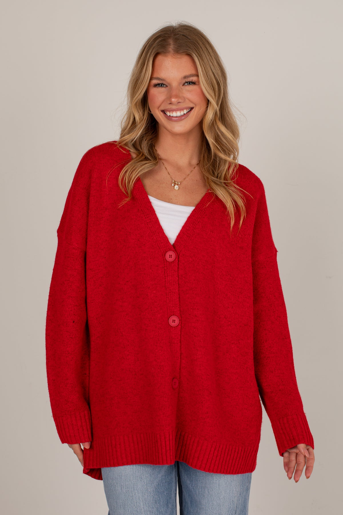 A smiling blonde woman wearing a bright red knit cardigan with three front buttons over a white top and light blue jeans, photographed against a plain light beige studio background.
