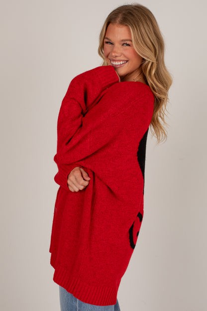A woman modeling a chunky knit red sweater with black contrast panels and a large black letter detail, photographed against a plain light gray studio background.