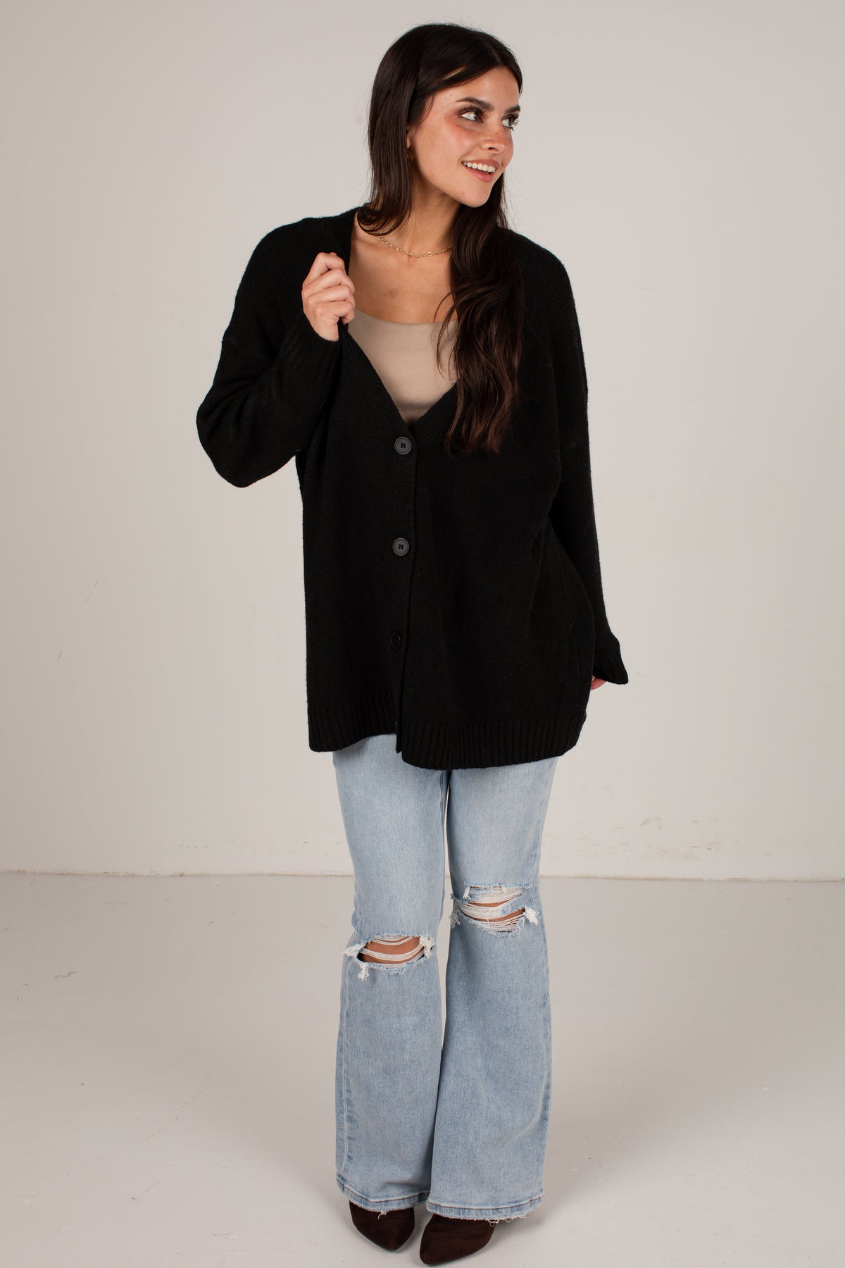 A woman modeling a black button-up knit cardigan over a beige top and light-wash distressed flare jeans, standing against a plain light gray studio background.