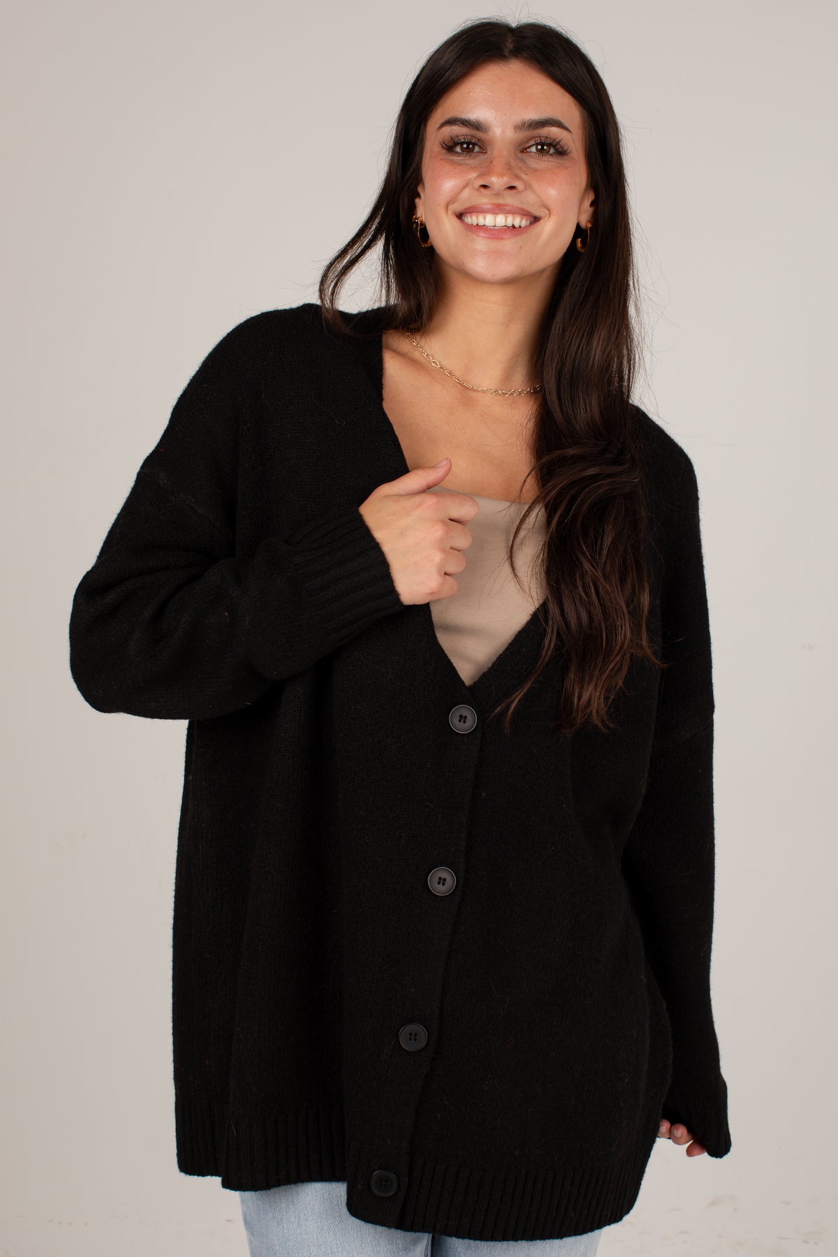 A woman wearing a black knit cardigan sweater over a beige top, standing against a plain light gray background.