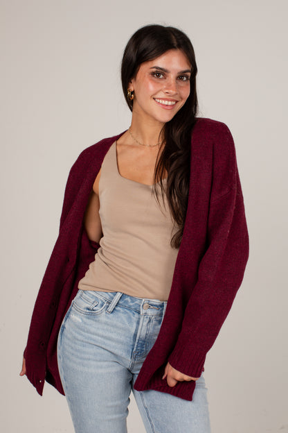 A woman modeling a heathered burgundy open-front knit cardigan over a beige tank top and light-wash blue jeans, photographed against a plain light gray studio background.