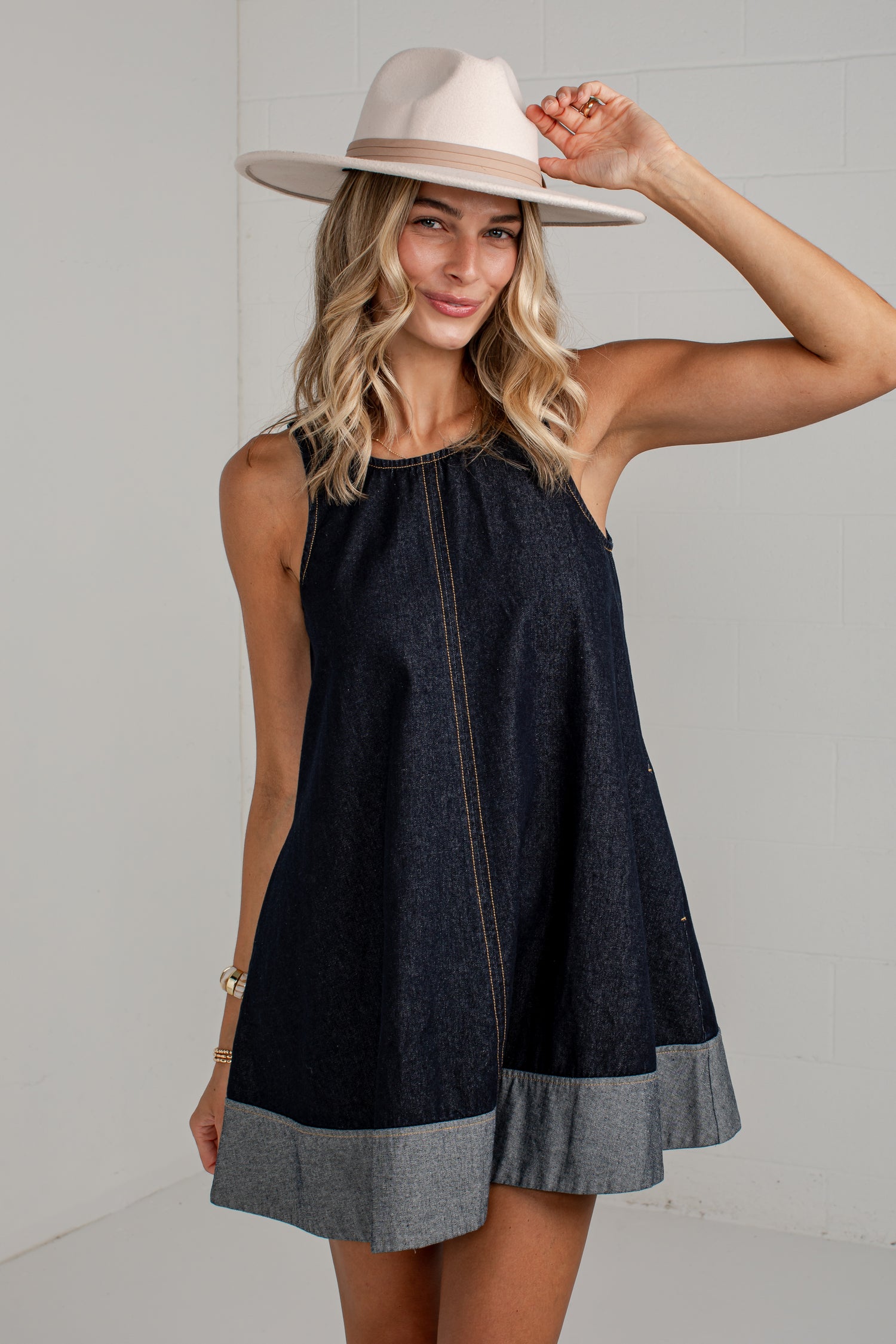 A woman wearing a sleeveless dark denim mini dress with a contrasting heather gray ruffle hem and a light beige wide-brim hat, posing against a white brick wall.