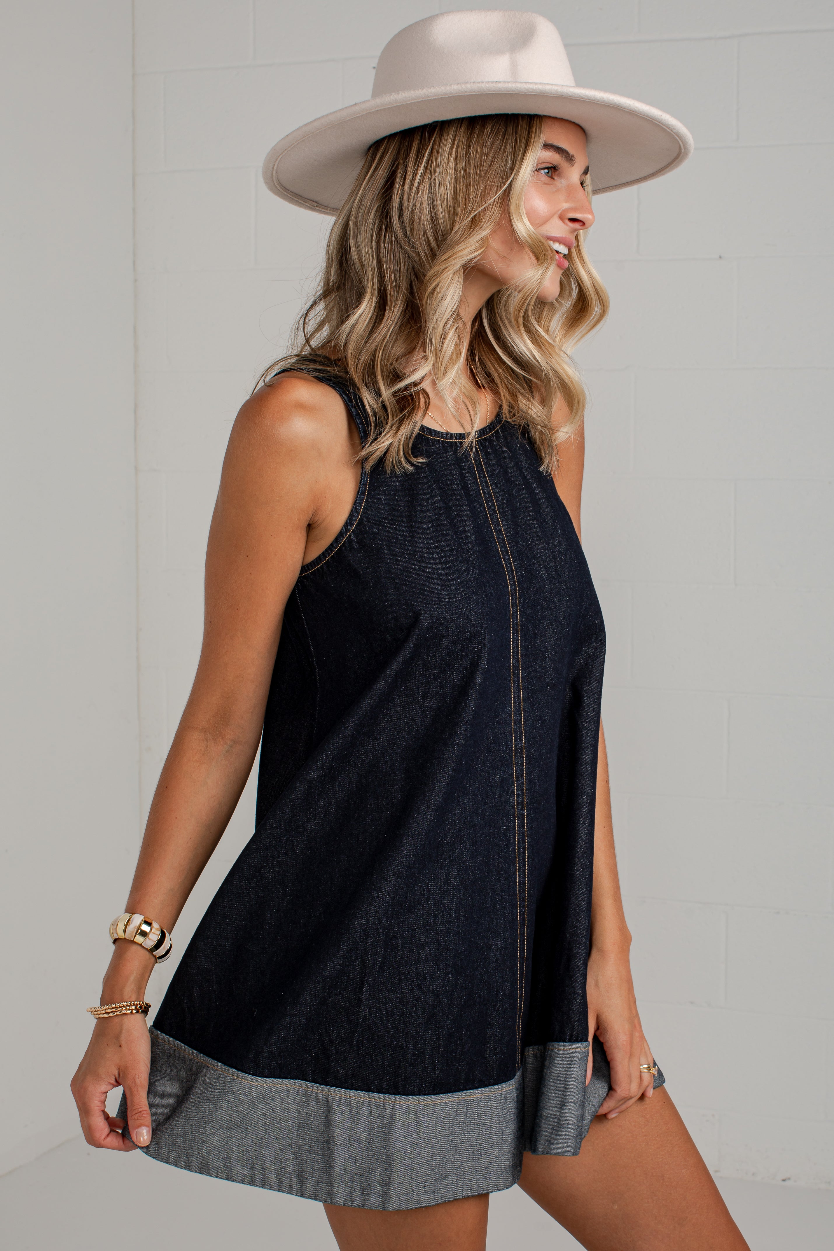 A woman wearing a sleeveless dark denim mini dress with a contrasting light gray hem and a wide-brimmed beige hat, posing against a light gray brick wall.