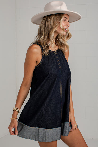 A woman wearing a sleeveless dark denim mini dress with a contrasting light gray hem and a wide-brimmed beige hat, posing against a light gray brick wall.