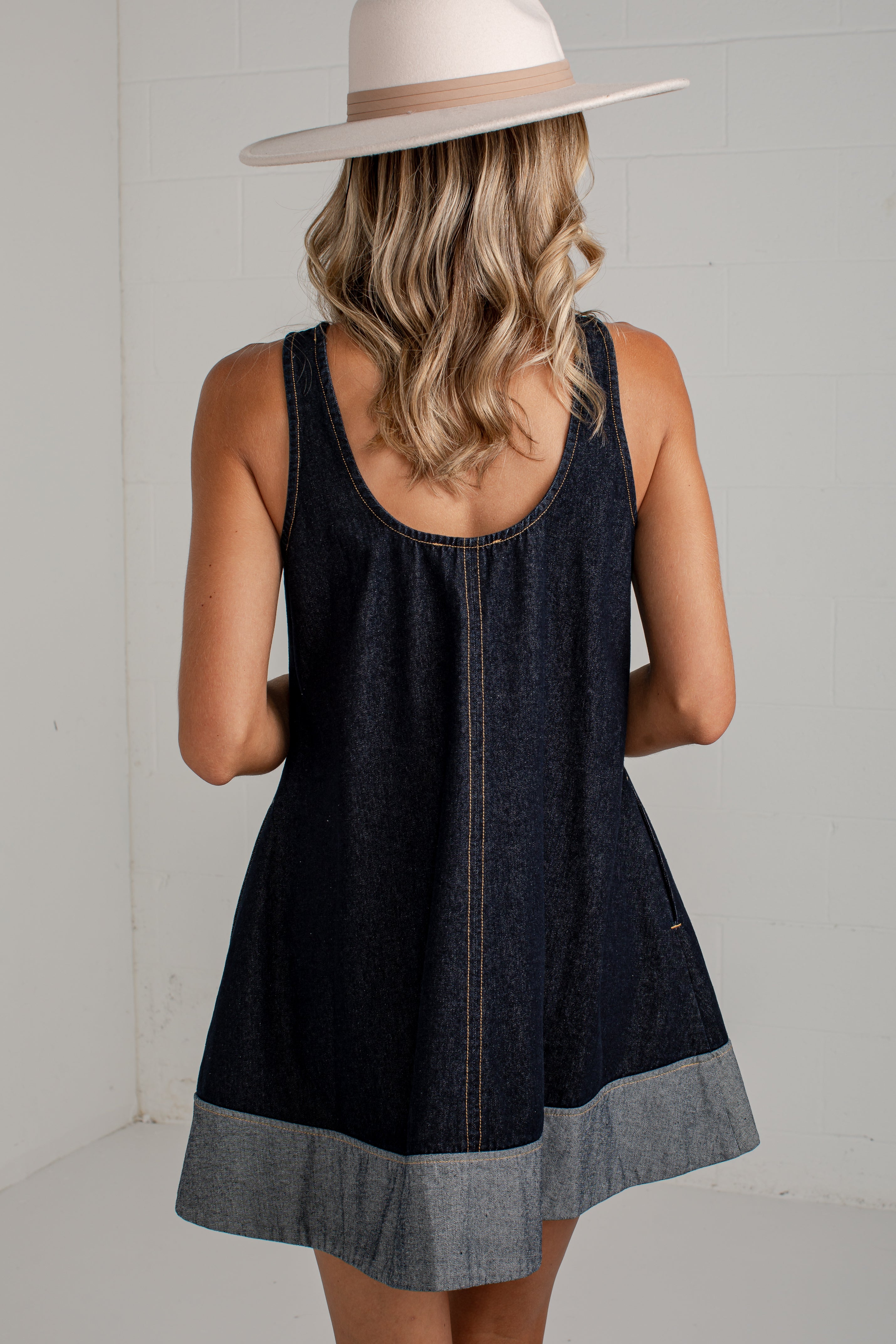 A woman viewed from behind wearing a sleeveless dark denim dress with a lighter gray cuffed hem and a wide-brim beige hat, standing against a white wall.