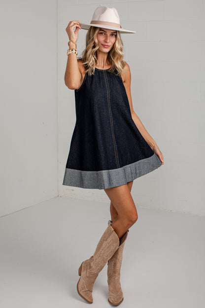 A woman modeling a sleeveless dark denim dress with a lighter gray hem, styled with a cream wide-brim hat and tan cowboy boots against a white studio background.