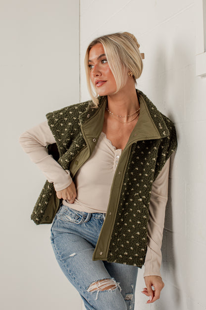 A woman wearing an olive green zip-front vest with a white floral pattern over a beige long-sleeve top and blue jeans, posing against a white wall.