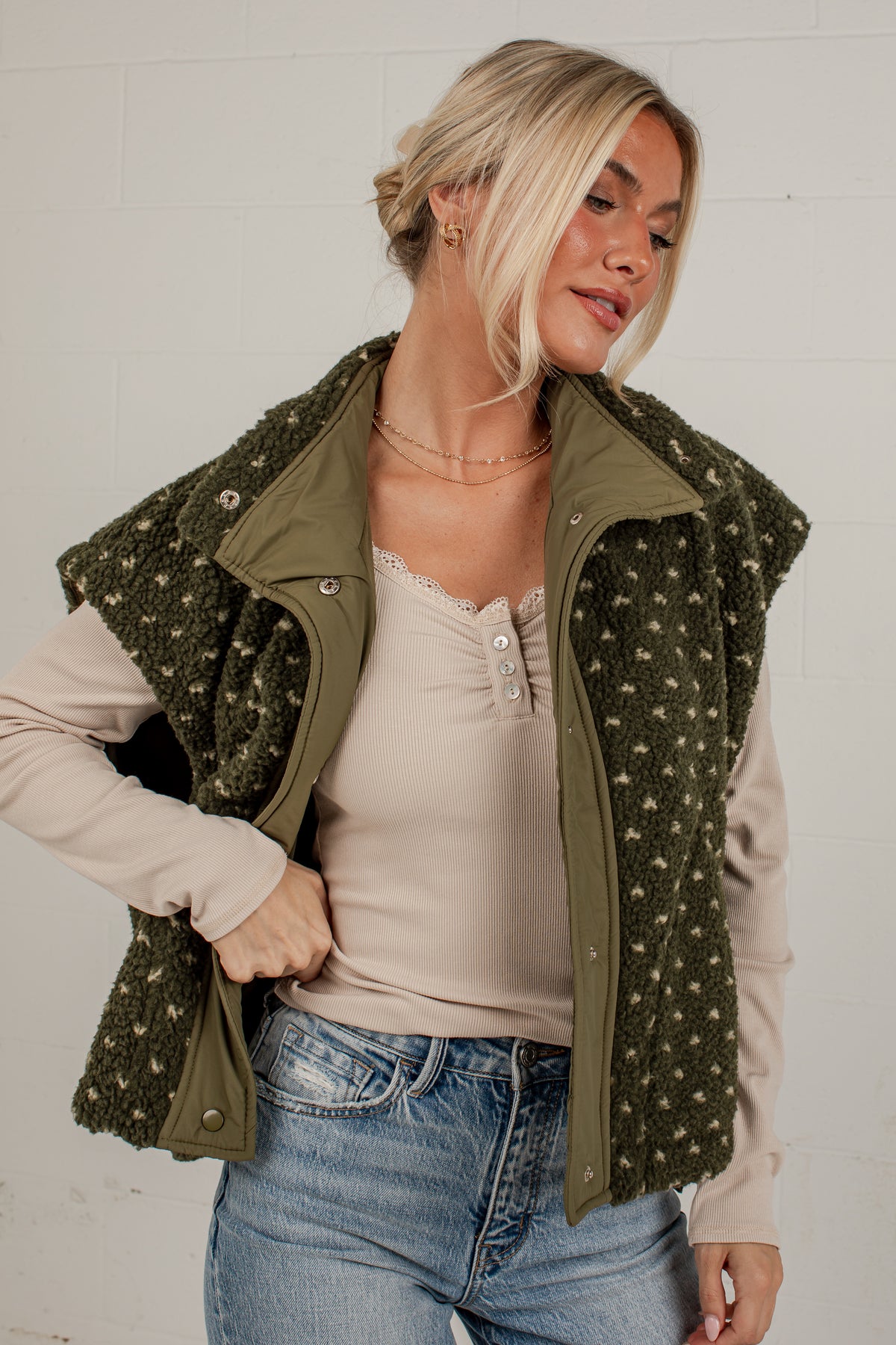 A woman wearing an olive green textured vest over a beige henley top and blue jeans, standing against a white brick wall.