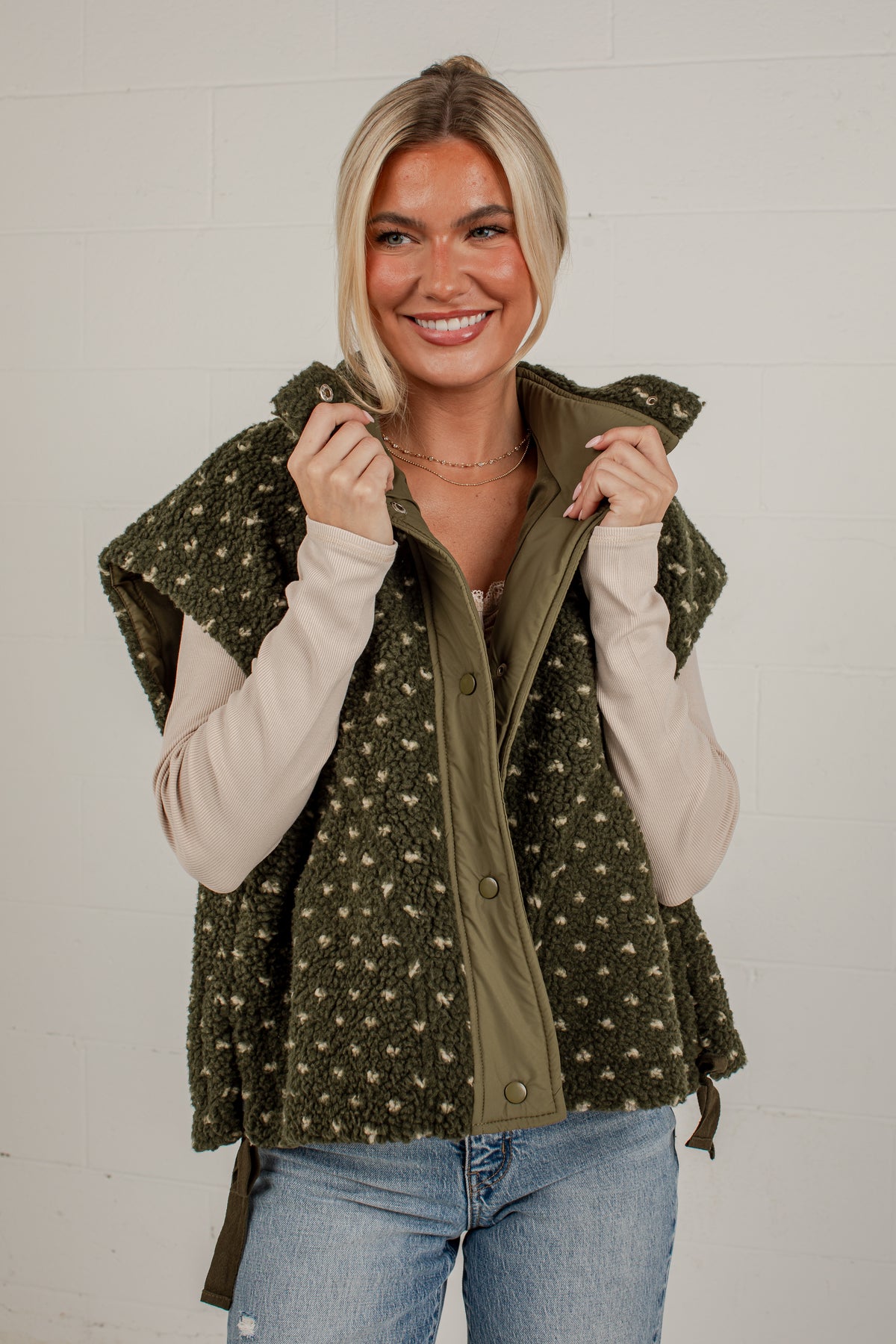 A smiling blonde woman wearing a sleeveless olive green vest with a beige and green pattern over a cream long-sleeve shirt and blue jeans, standing against a white brick wall.