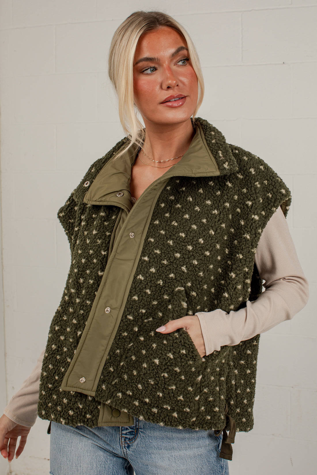 A woman wearing an olive green fleece vest with a beige and white speckled pattern, layered over a beige long-sleeve shirt and blue jeans, standing against a white brick wall.