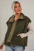 A woman wearing an olive green fleece vest with a beige and white speckled pattern, layered over a beige long-sleeve shirt and blue jeans, standing against a white brick wall.