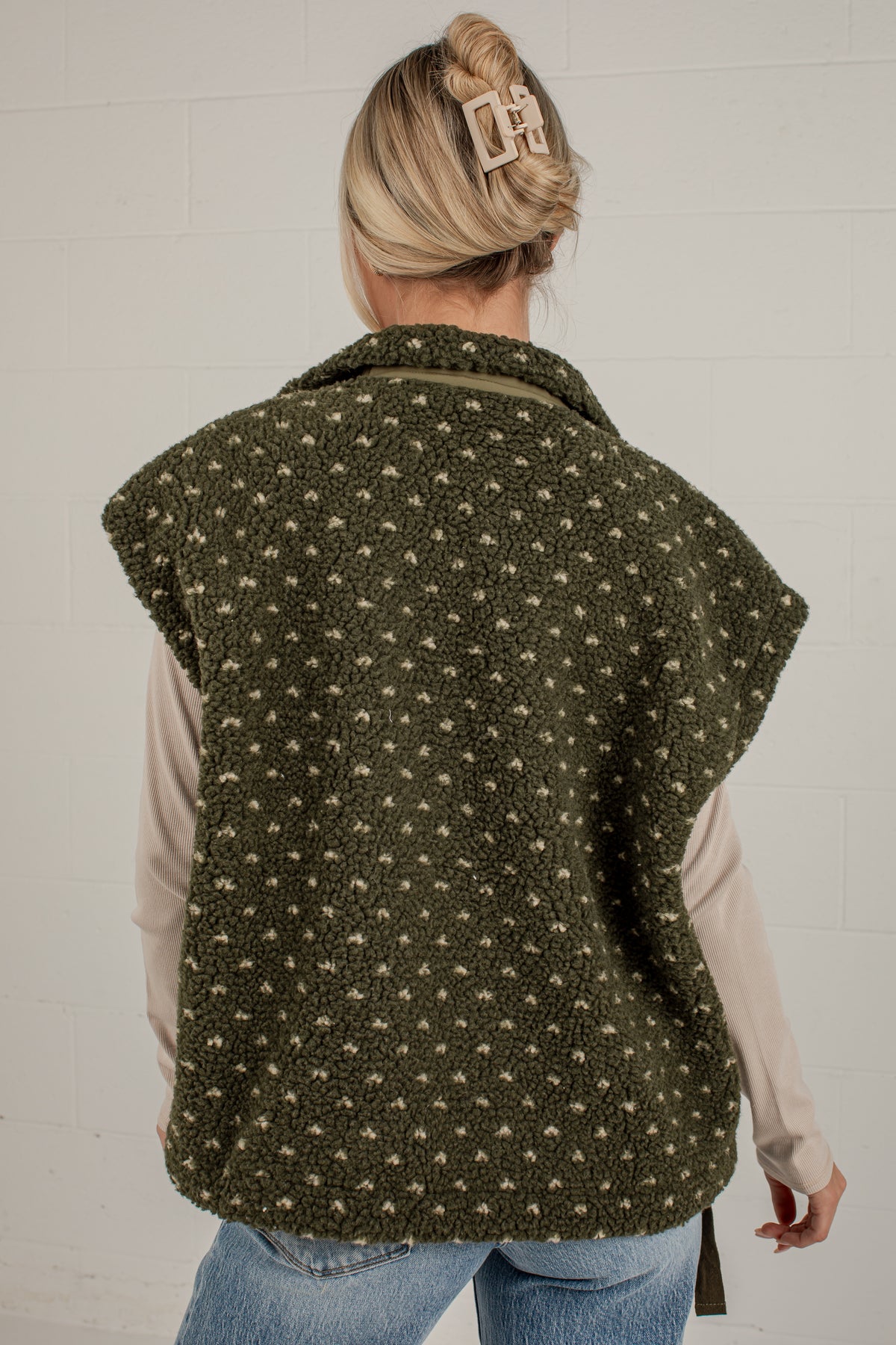 A woman shown from the back wearing a dark olive green fleece vest with a small beige geometric pattern, layered over a light beige long-sleeve shirt and blue jeans against a white brick wall.