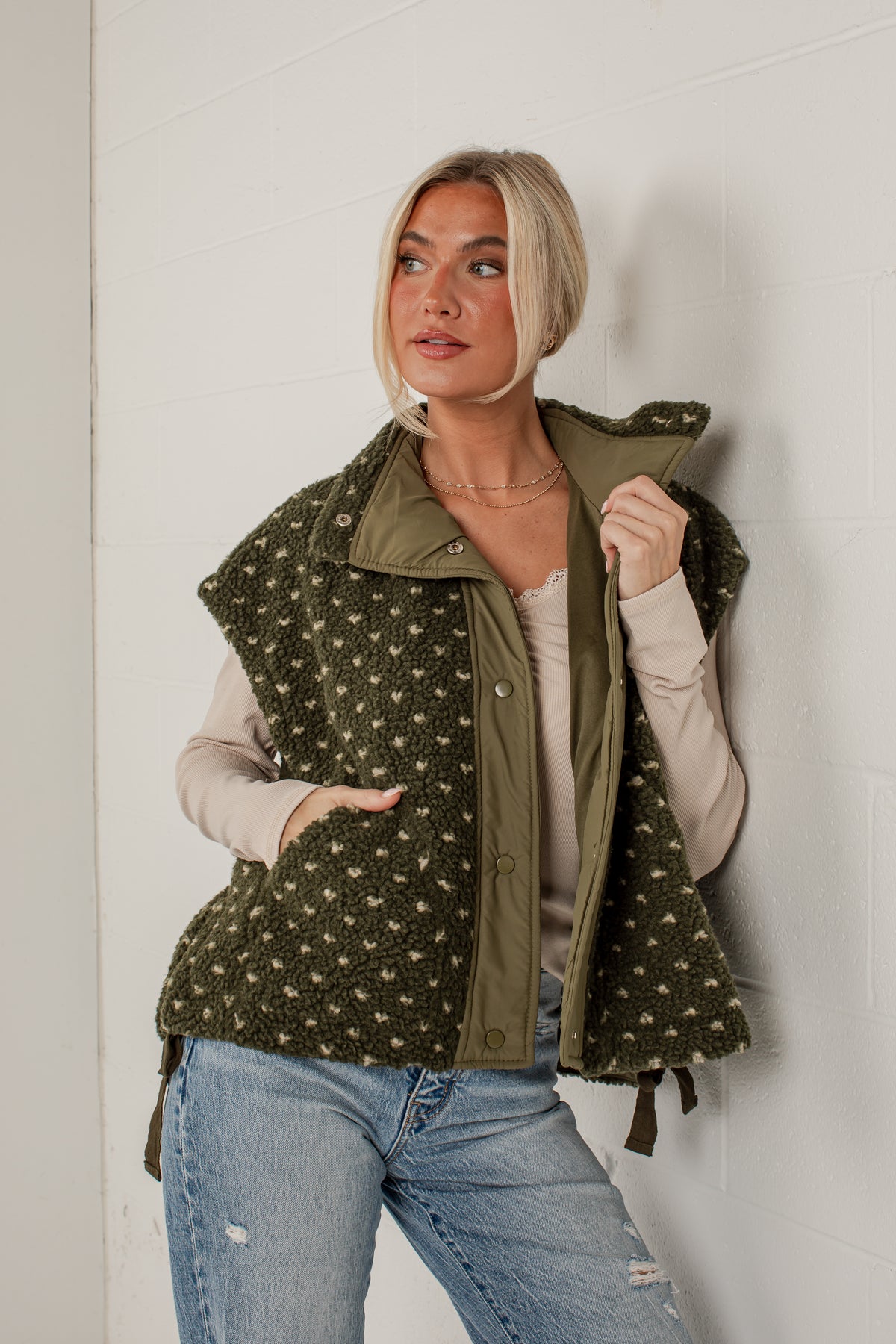A woman wearing an olive green fleece vest with a beige and green floral pattern, styled over a beige top and blue jeans, leaning against a white brick wall.