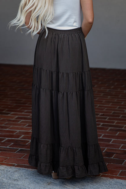 A woman wearing a long, tiered black skirt with a white sleeveless top, standing outdoors on a brick surface.