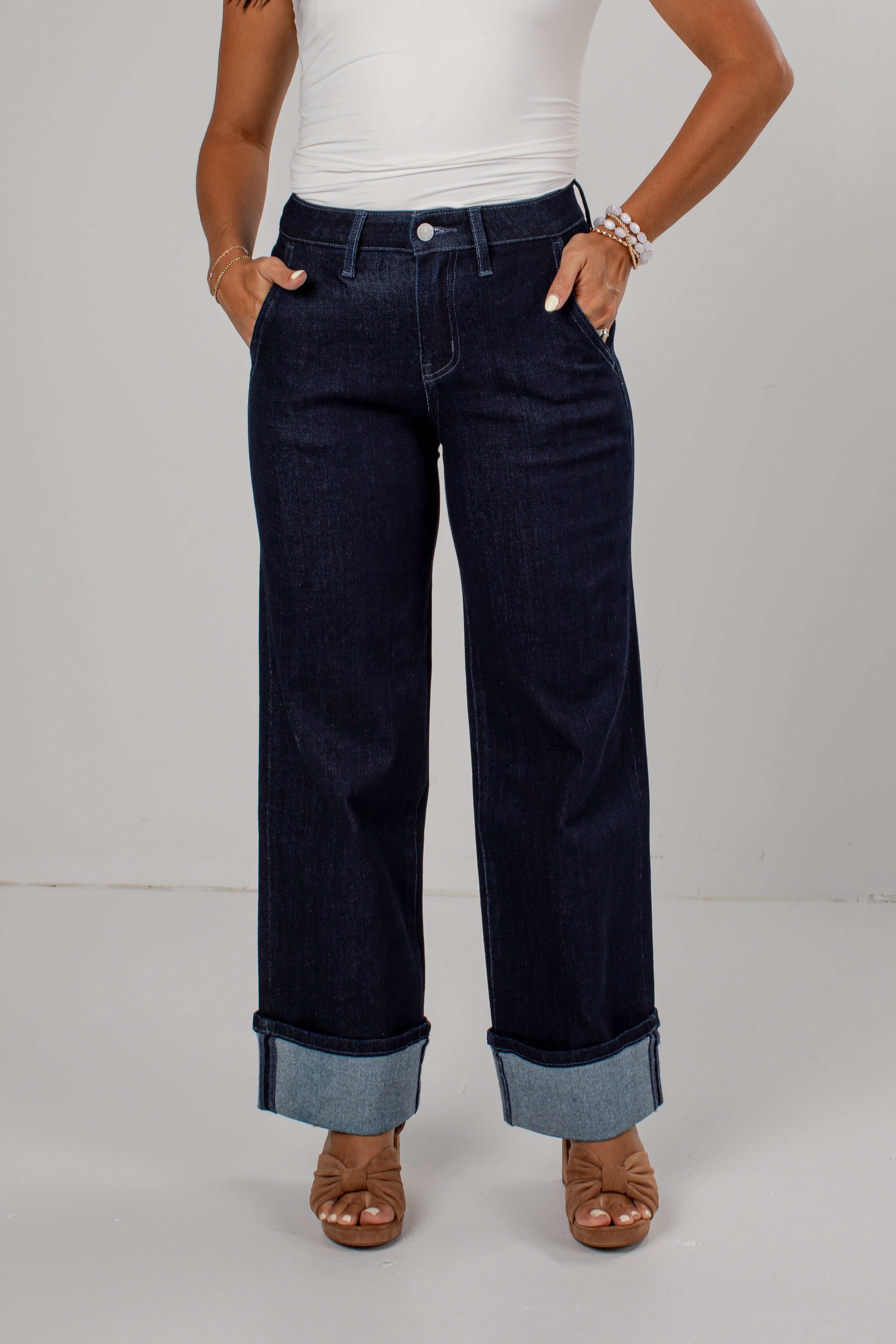A model wearing dark wash wide-leg jeans with rolled cuffs, paired with a white top and brown sandals, standing against a light gray studio background.