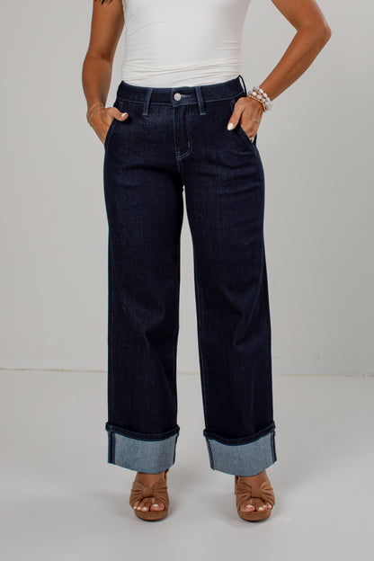 A model wearing dark wash wide-leg jeans with rolled cuffs, paired with a white top and brown sandals, standing against a light gray studio background.