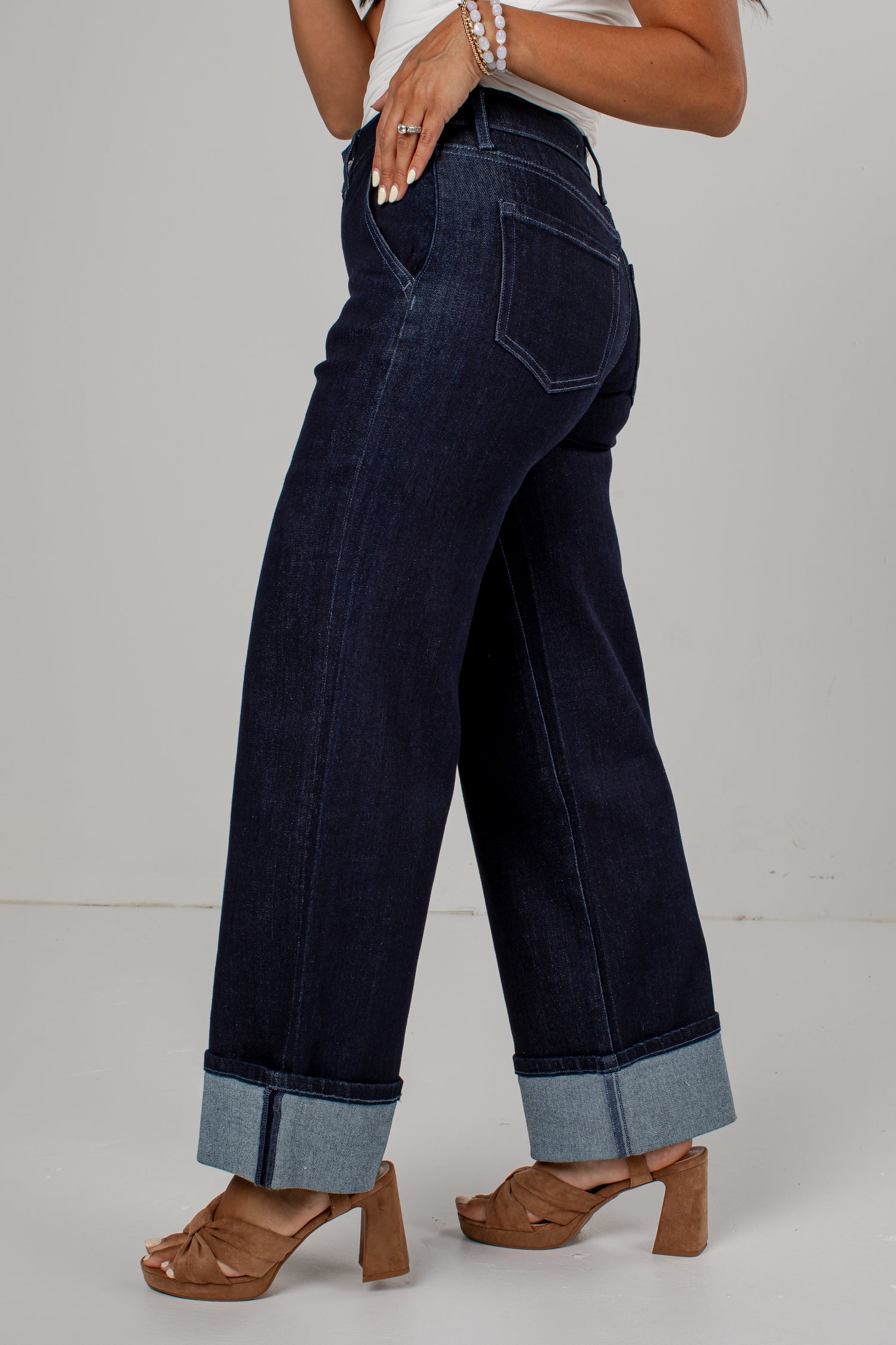 A person wearing dark blue wide-leg cuffed jeans with rolled cuffs, paired with brown heeled sandals and a white top, shown from the waist down against a light gray studio background.