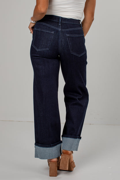 Rear view of a person wearing dark indigo denim jeans with rolled cuffs, paired with a white top and brown block-heel sandals, against a light gray studio background.