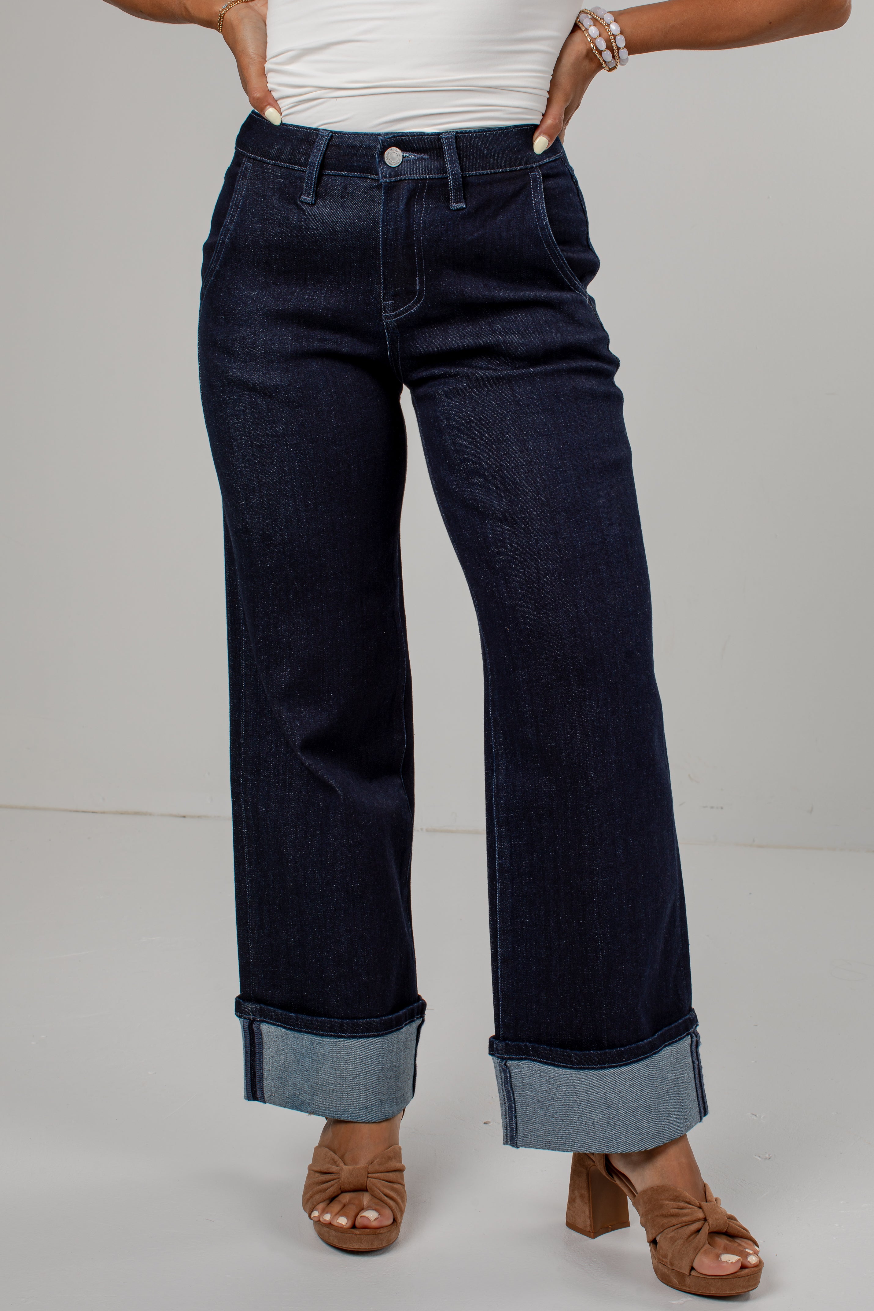 A person wearing dark indigo wide-leg jeans with rolled cuffs, paired with a white top and brown heeled sandals, shown from the waist down against a light gray studio background.