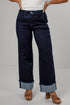 A person wearing dark indigo wide-leg jeans with rolled cuffs, paired with a white top and brown heeled sandals, shown from the waist down against a light gray studio background.