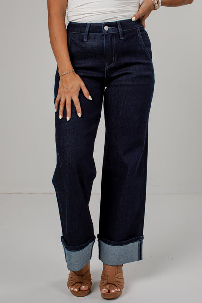 A person wearing dark indigo blue wide-leg jeans with rolled cuffs, paired with a white top and brown open-toe heeled sandals, shown from the waist down against a light gray studio background.