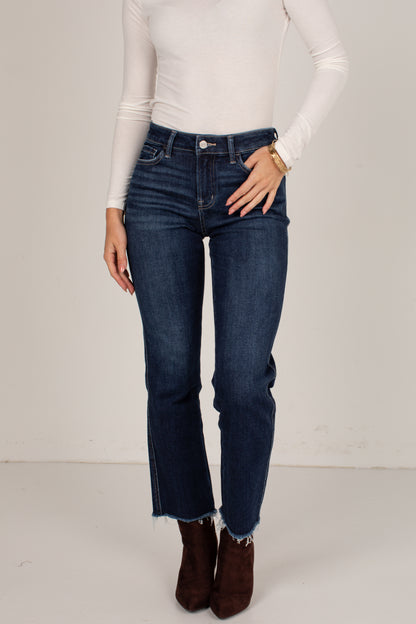 A model wearing dark blue high-waisted straight-leg jeans with a raw frayed hem, styled with a white long-sleeve top and brown suede ankle boots against a plain off-white studio background.