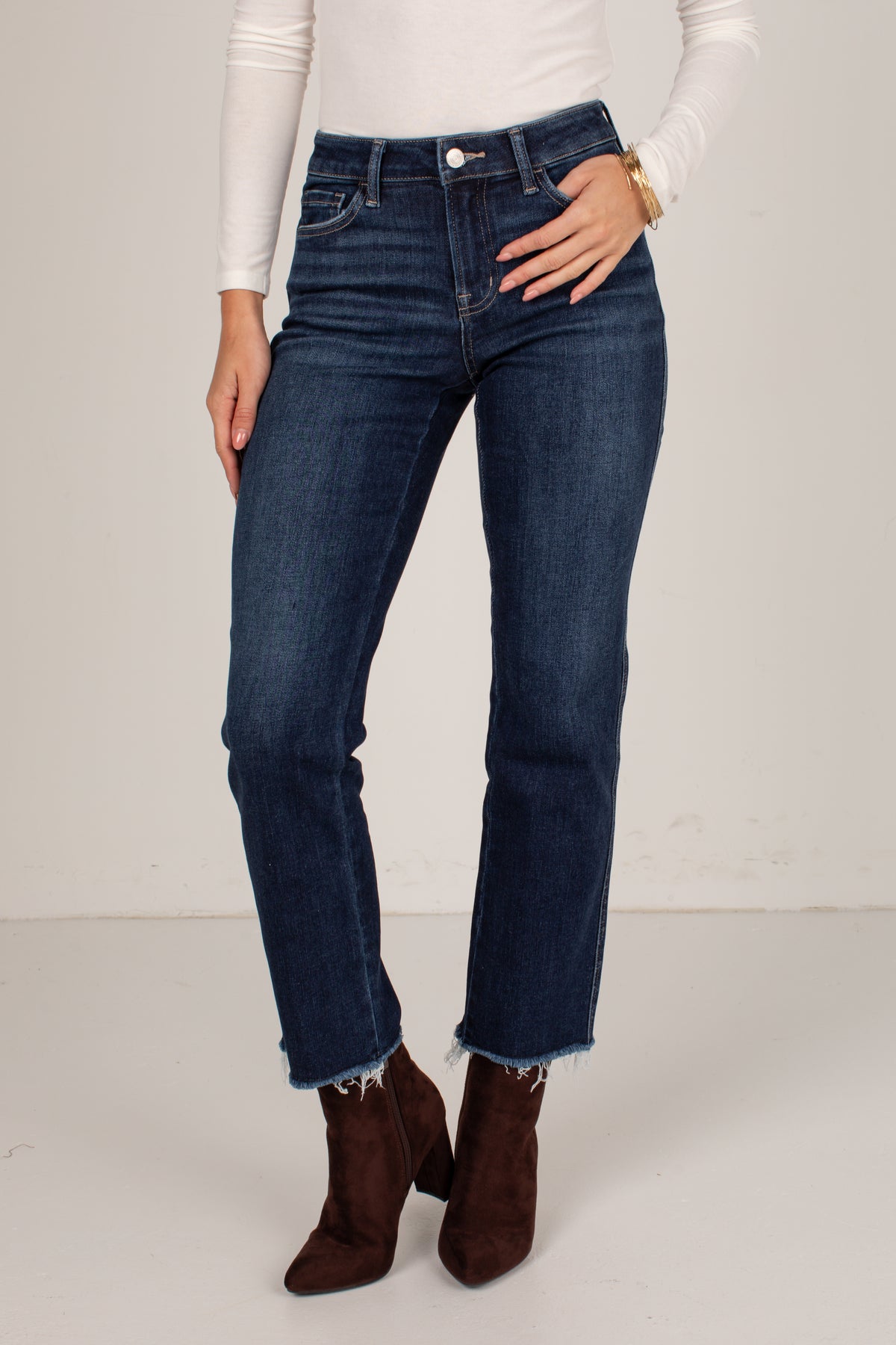 A model wearing dark blue straight-leg denim jeans with a raw, frayed hem, styled with a white long-sleeve top and dark brown suede ankle boots against a plain off-white studio background.