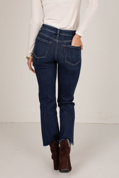 A person shown from the back wearing dark blue denim jeans with a raw frayed hem, paired with a white long-sleeve top and brown ankle boots, against a plain off-white studio background.