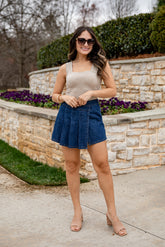 Woman wearing a beige tank top and blue denim skirt standing outdoors with stone wall and flowers in the background.