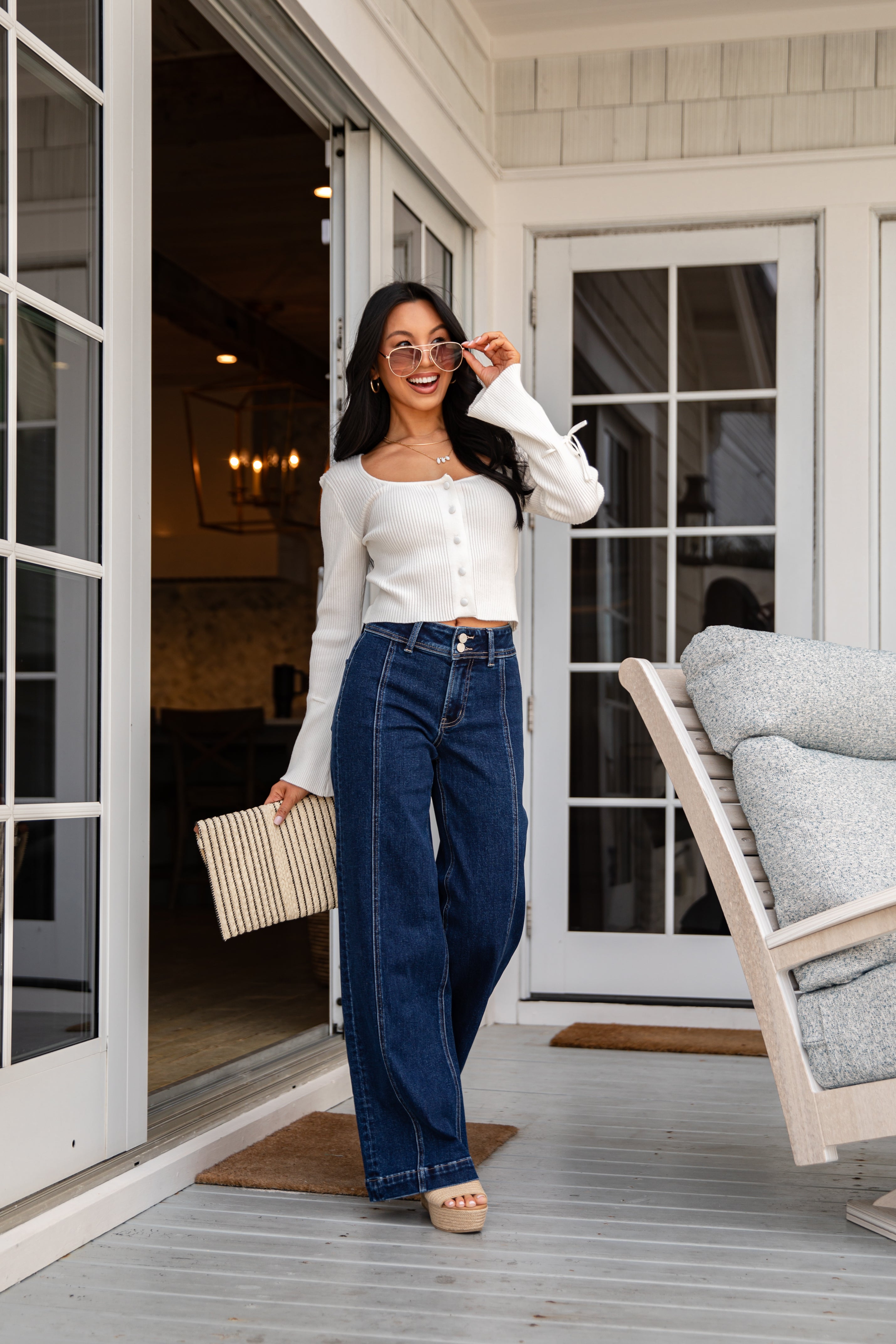 Woman standing on a porch wearing a white blouse and blue jeans, holding a beige clutch.