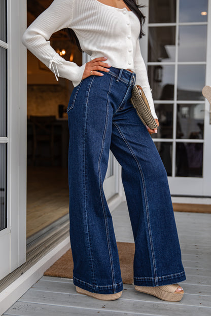 Person wearing a white top and blue jeans standing on a wooden deck.