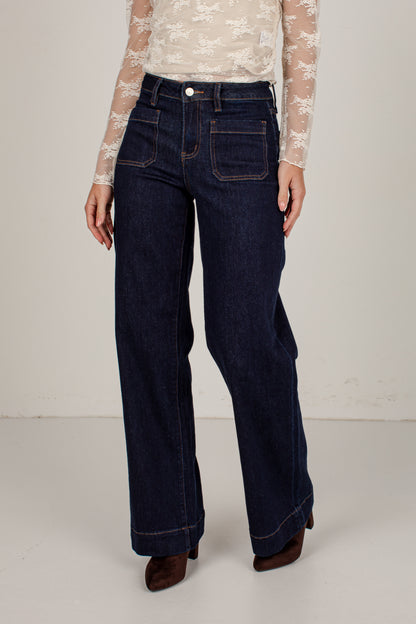 A person wearing dark blue wide-leg denim jeans with a cream lace long-sleeve top and brown pointed-toe heels, standing against a plain light gray studio background.