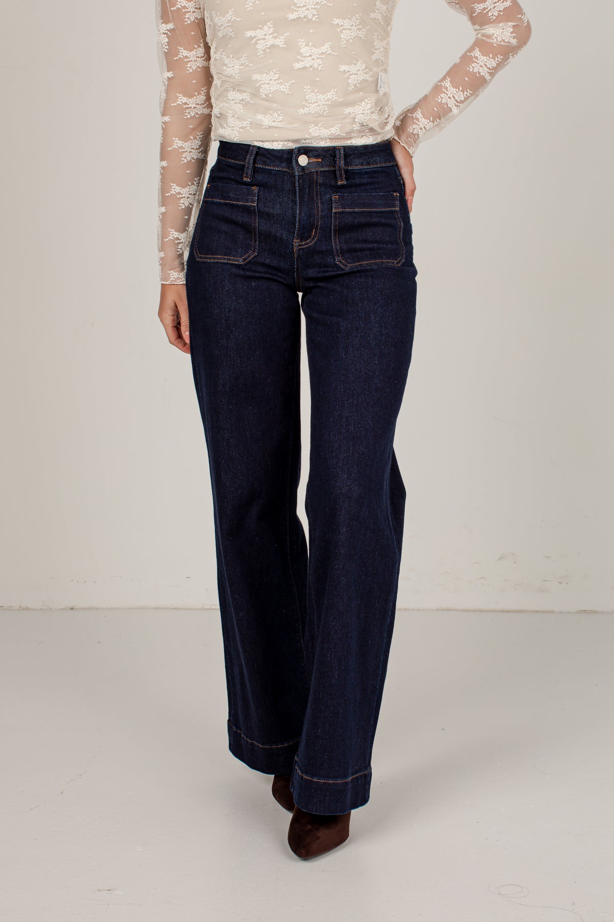 A model wearing dark blue high-waisted flare jeans, paired with a cream lace long-sleeve top and brown pointed-toe heels, standing against a plain off-white studio background.