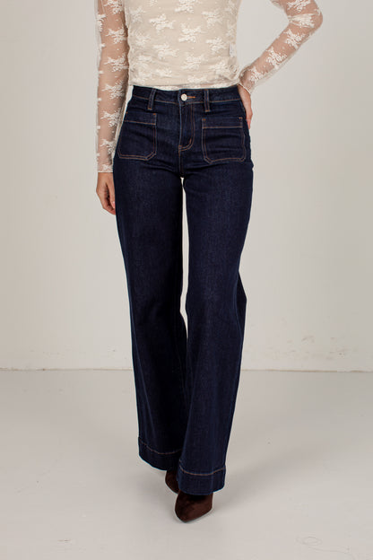 A model wearing dark blue high-waisted flare jeans, paired with a cream lace long-sleeve top and brown pointed-toe heels, standing against a plain off-white studio background.