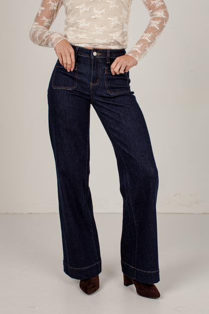A model wearing dark blue wide-leg denim jeans with a cream lace long-sleeve top and brown pointed-toe heels, standing against a plain off-white studio background.