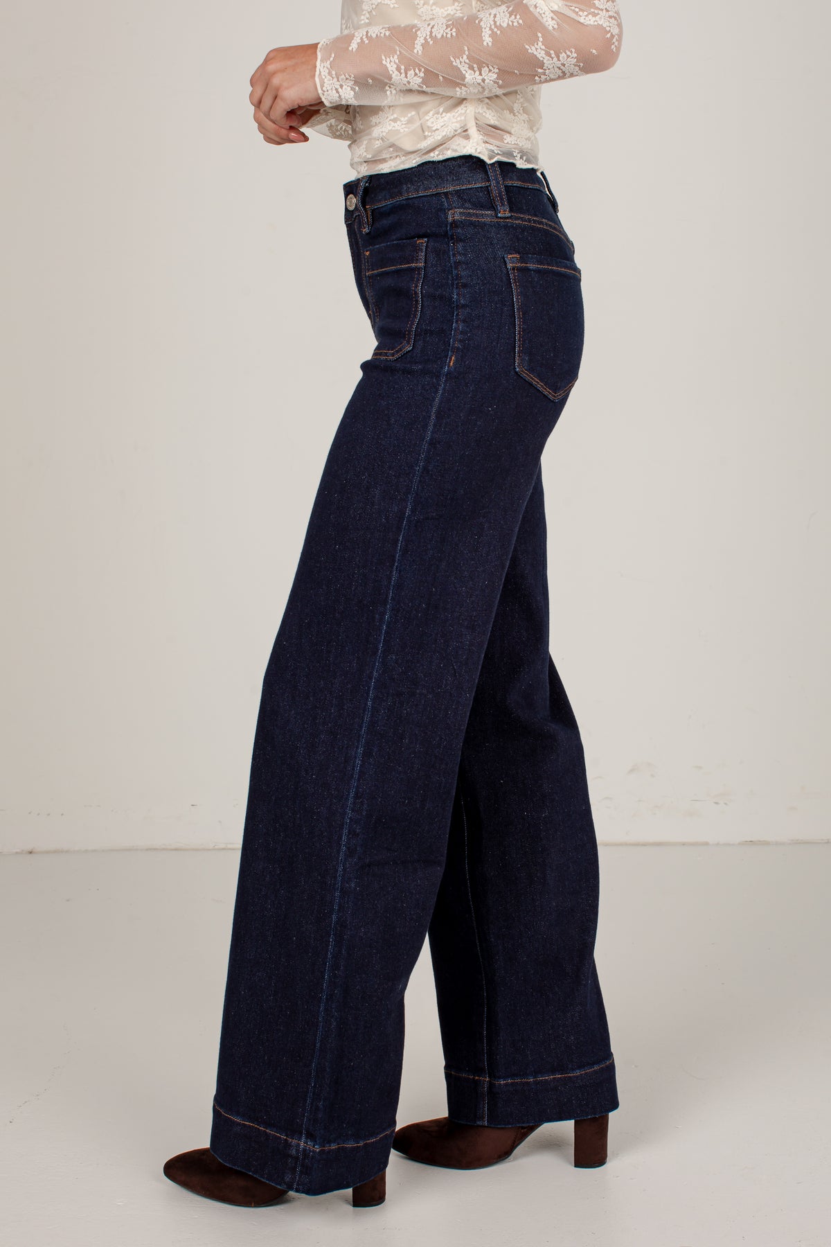 A person shown from the waist down wearing dark blue wide-leg jeans, a white lace long-sleeve top, and brown heeled shoes against a plain off-white studio background.